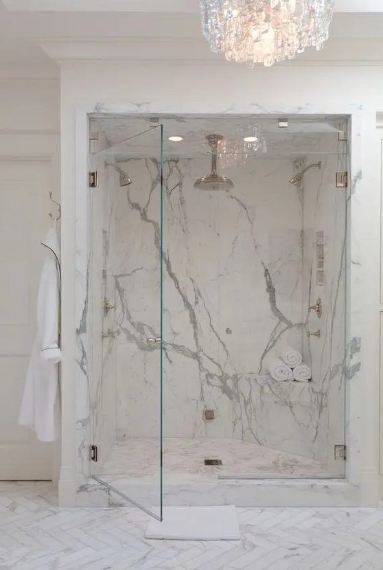 a luxurious bathroom with a walk-in shower clad with white marble tiles and with glass door plus a refined crystal chandelier