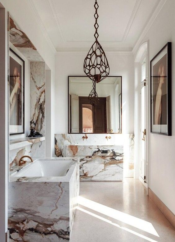 a luxurious modern French bathroom with a marble wall and a tub clad with it, a marble vanity, black metal pendant lamps and brass fixtures