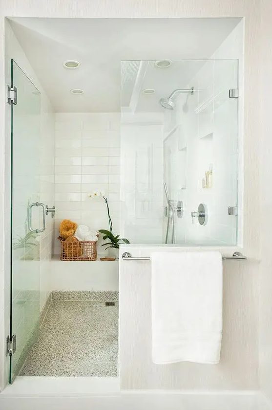 a luxurious modern white shower space with glass doors, white tiles and a terrazzo floor, a basket for storage and an orchid