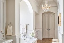 a luxury neutral French bathroom with neutral furniture, a tub clad with white marble, crystal chandeliers and a neutral vintage chair