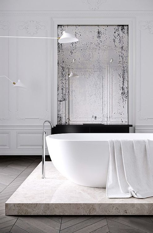 a minimalist French chic bathroom with a tub on a stone platform, a vintage mirror, stucco walls and catchy minimal lamps