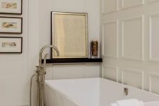 a modern French chic bathroom in neutrals with paneling, a square tub, a ledge with art and a small gallery wall for more elegance