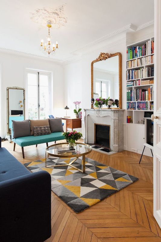 a modern and chic French living room with a marble fireplace, a bold turquoise loveseat and a navy sofa, mirrors and a glass coffee table