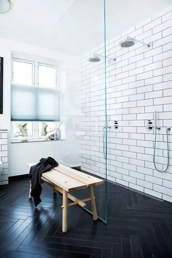 a modern bathroom with a black herringbone floor, white subway tiles in the walk in shower, with a glass paritition and a wooden bench