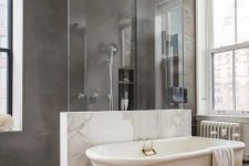 a modern bathroom with concrete walls and a floor, with a half wall and glass partition in the shower, a vintage tub