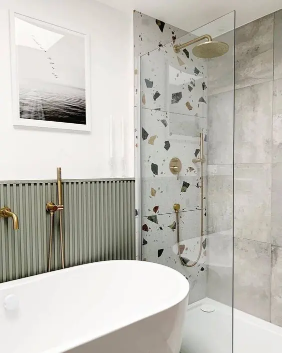 a modern bathroom with green paneling, a walk in shower with concrete and a terrazzo wall, brass fixtures and a glass partition