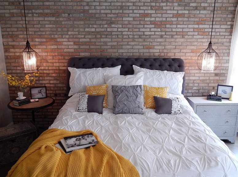 a modern industrial bedroom with a faux brick wall, a grey upholstered bed, a round table and an inlay dresser, cool pendant lamps