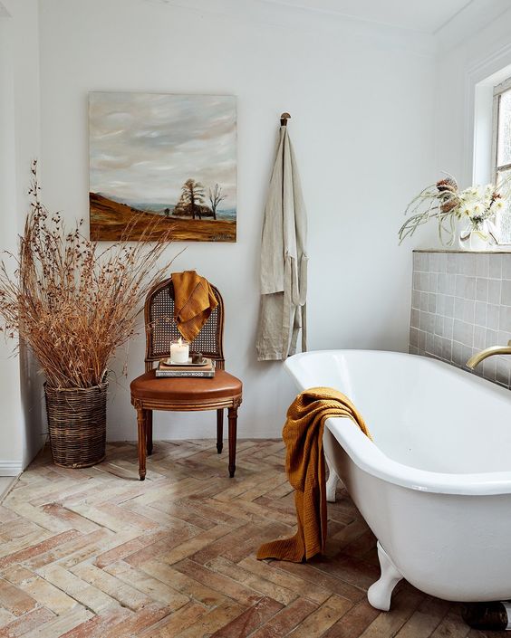 a neutral French bathroom with a grey tile wall, a herringbone floor, a clawfoot tub, a leather chair and dried branches in a basket