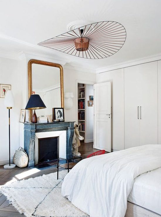 A neutral Parisian bedroom with a blue non-working fireplace, a statement lamp, a chic mirror and an antique wooden chair