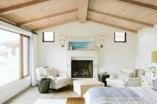a neutral and beautiful bedroom with a panoramic window, a blonde wood ceiling with beams, a fireplace, white chairs and an ottoman, a bed and leather stools