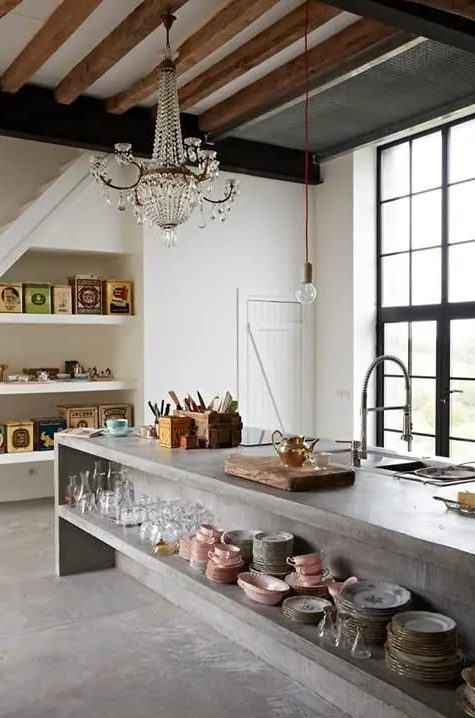 a neutral kitchen with wooden beams on the ceiling a large concrete kitchen island with long open shelves and built in ones on one of the walls