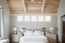 a pretty modern bedroom with a wooden ceiling and beams, a grey upholstered bed with neutral bedding, mismatching sideboards as nightstands and a series of windows