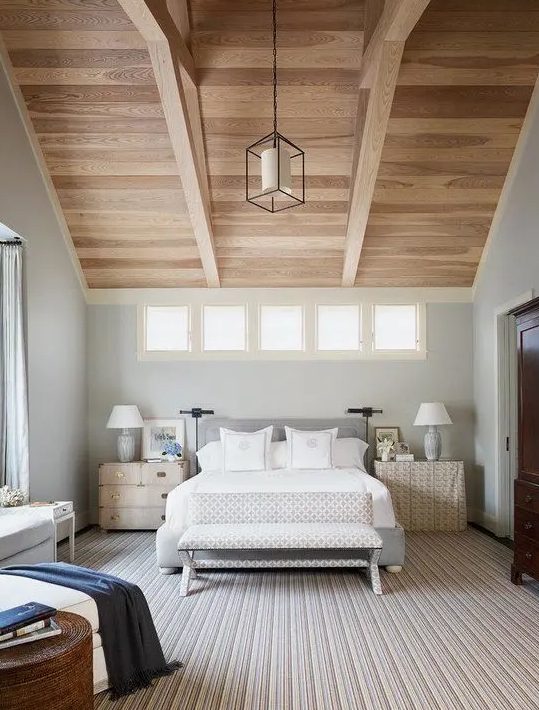 a pretty modern bedroom with a wooden ceiling and beams, a grey upholstered bed with neutral bedding, mismatching sideboards as nightstands and a series of windows
