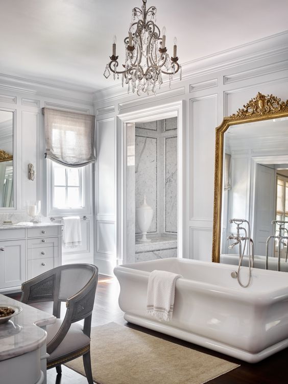 a refined French style bathroom in white, with a vanity, a tub and a mirror in a gilded ornated frame, a crystal chandelier and blinds on the curtains