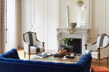 a refined French style living room with a non-working fireplace, a bold blue sofa, striped vintage chairs, a chic crystal chandelier and a clear acrylic table