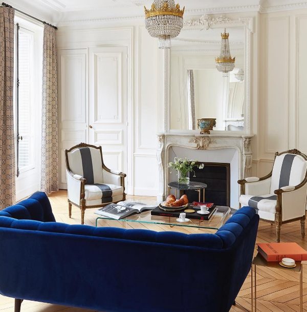 a refined French style living room with a non working fireplace, a bold blue sofa, striped vintage chairs, a chic crystal chandelier and a clear acrylic table