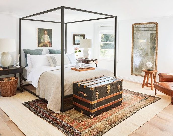 a refined bedroom with a black frame bed with neutral bedding, mismatching yet similar-looking dark stained nightstands, a black chest for storage and some artworks