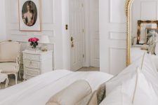 a refined white Parisian bedroom with paneling, a white bed, an oversized mirror, some chic vintage furniture and a beautiful artwork