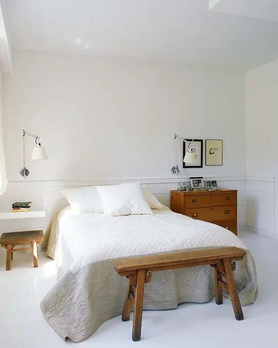 a serene white bedroom with paneling, a bed with a bench, a floating nightstand with a stool and a stained dresser as a nightstand