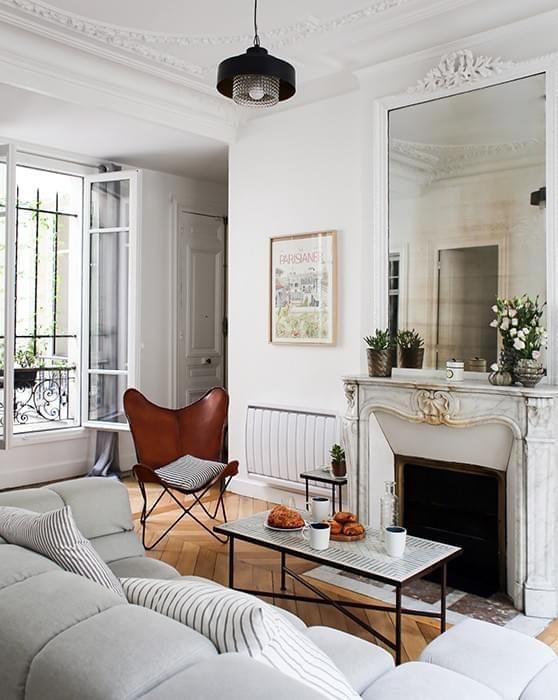 a small yet chic French style living room with an antique fireplace, a coffee table, a brown butterfly chair and a grey sofa