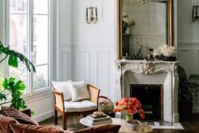 a sophisticated French chic living room with an antique fireplace, a large mirror in an ornated frame, a neutral chair, a rust-colored sofa with pillows and potted plants