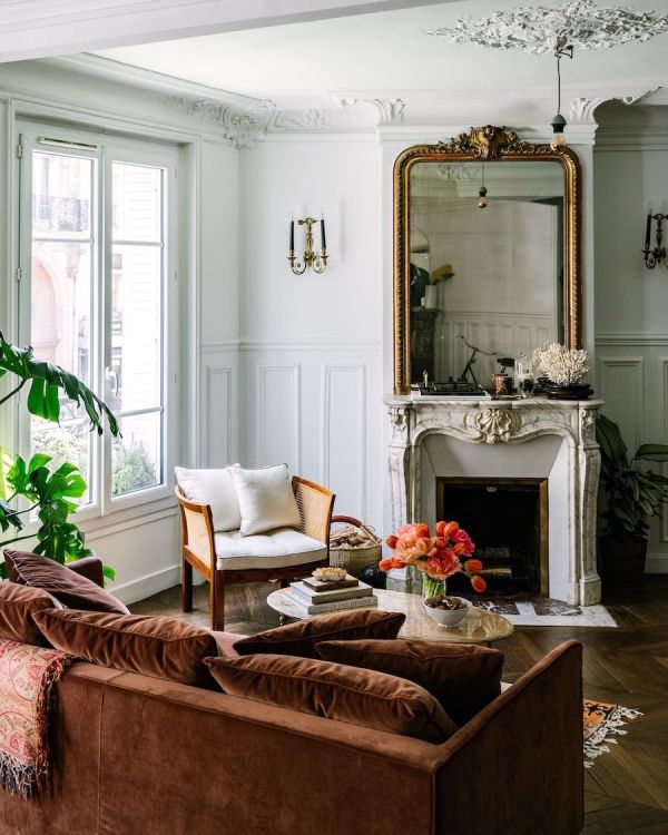 a sophisticated French chic living room with an antique fireplace, a large mirror in an ornated frame, a neutral chair, a rust-colored sofa with pillows and potted plants
