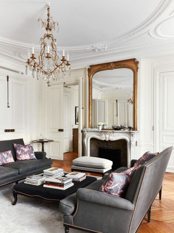 a sophisticated French style living room with an antique fireplace, graphite grey sofas, an elegant black ottoman and a crystal chandelier
