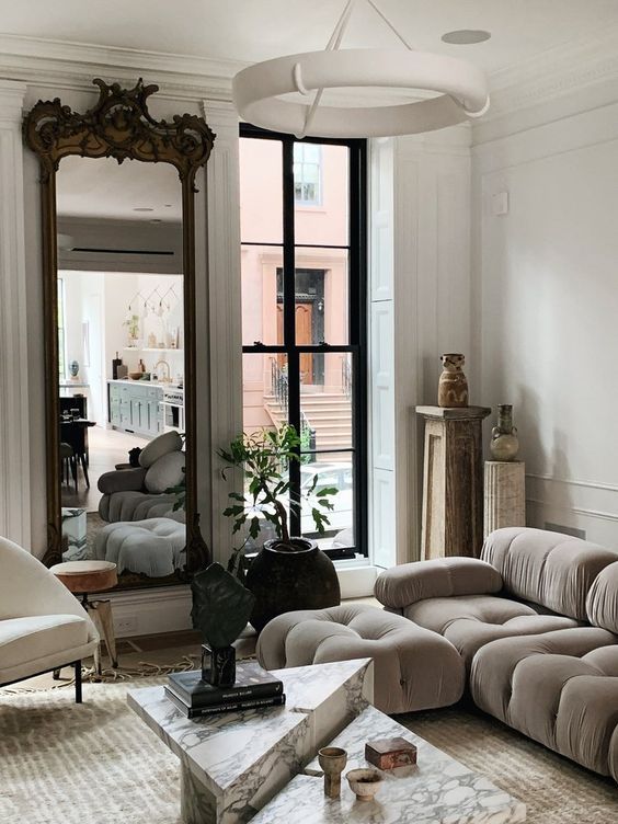 a sophisticated French style lviing room with a mirror in an ornated frame, a grey low sofa, a creamy chair and marble slab tables is jaw-dropping