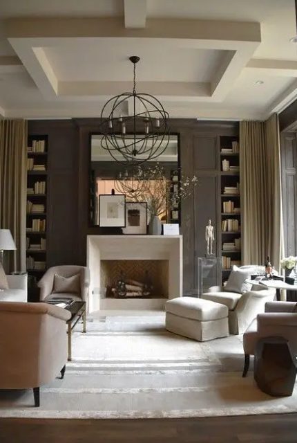 a sophisticated taupe living room with paneling, built-in bookshelves, a non-working fireplace, neutral seating furniture, a sphere pendant lamp and a large mirror
