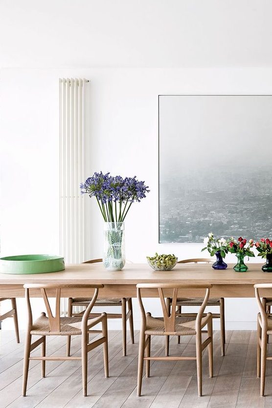 a vivacious dining room with blonde wood furniture, a beautiful artwork on the wall, blooms and greenery