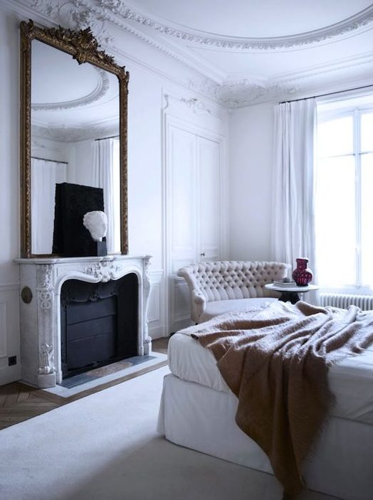 a white Parisian bedroom with a statement mirror over the fireplace, an upholstered bed, a chic tufted sofa and much natural light