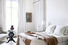 a white Parisian bedroom with airy curtains, an upholstered bed, an artwork and a chic tufted chair in the corner