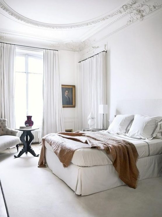 a white Parisian bedroom with airy curtains, an upholstered bed, an artwork and a chic tufted chair in the corner