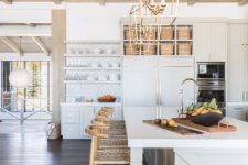 a white famrhouse kitchen with chic cabinets, a blonde wood ceiling with beams, woven stools, elegant brass pendant lamps
