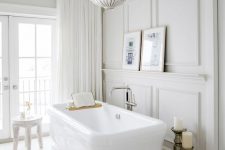 an airy and light-filled yet refined French bathroom with neutral paneling, a large tub, pillar candles, a chic chandelier and neutral curtains