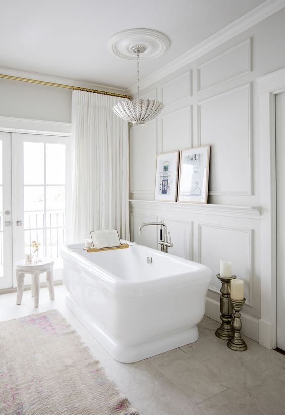 an airy and light filled yet refined French bathroom with neutral paneling, a large tub, pillar candles, a chic chandelier and neutral curtains