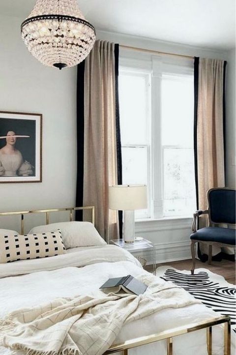 an elegant French chic bedroom with a gold bed with neutral bedding, a zebra printed rug, a navy chair, color block curtains and a lovely chandelier