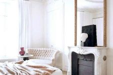 an exquisite bedroom with a French feel, with a non-working fireplace, a white bed with a tan blanket, a refined loveseat and an oversized mirror
