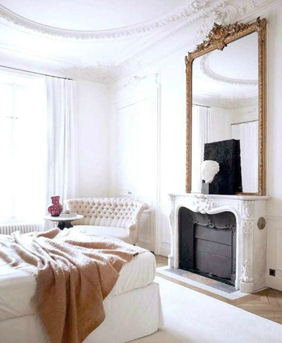 an exquisite bedroom with a French feel, with a non-working fireplace, a white bed with a tan blanket, a refined loveseat and an oversized mirror