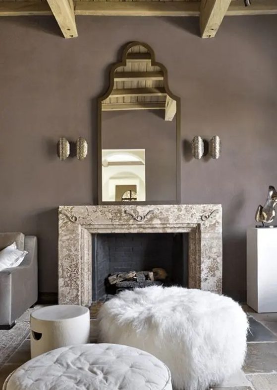 an exquisite taupe living room with a non working fireplace, a refined mirror, metallic sconces, white ottomans and poufs and taupe furniture