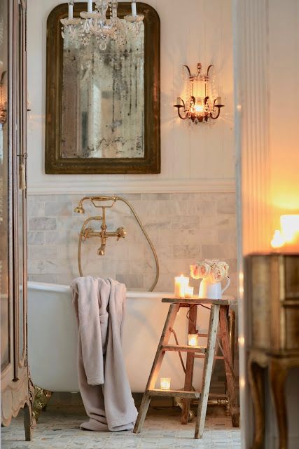 an inspiring French bathroom with a white marble tile wall, a vintage mirror, a clawfoot tub, pillars and crystal lamps and chandeliers