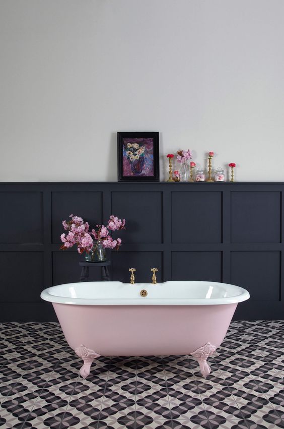 a beautiful bathroom with printed tiles on the floor, black paneling on the walls, a pink clawfoot bathtub, blooms and lovely artwork is amazing