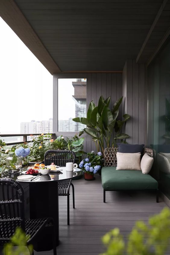 a stylish modern balcony with a black round table, black wicker chairs, a wicker daybed with some pillows, blooms and greenery around