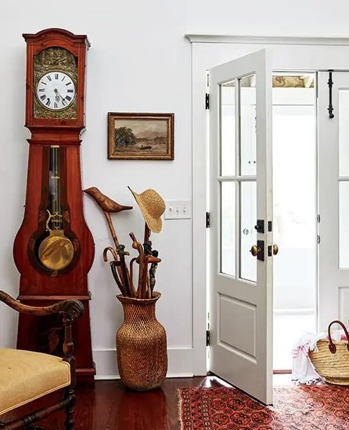 a chic entryway with a red wood clock, a wicker vase for umbrellas and a refined chair