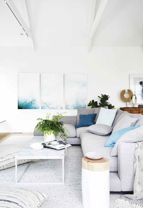 a beautiful ocean-inspired living room with an ocean-inspired three piece artwork, a grey sectional and pillows, a coffee table and lots of negative space for an airy feel
