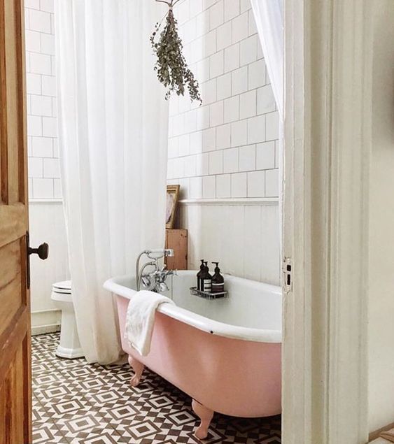a chic bathroom with black and white tiles on the floor, white square tiles on the walls, a pink clawfoot bathtub, some greenery and white curtains
