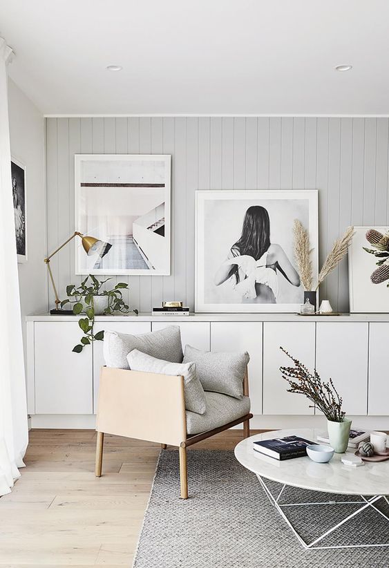 a neutral Scandinavian living room with a grey accent wall, a plain storage unit, a gallery wall, a grey chair, a round coffee table and some greenery
