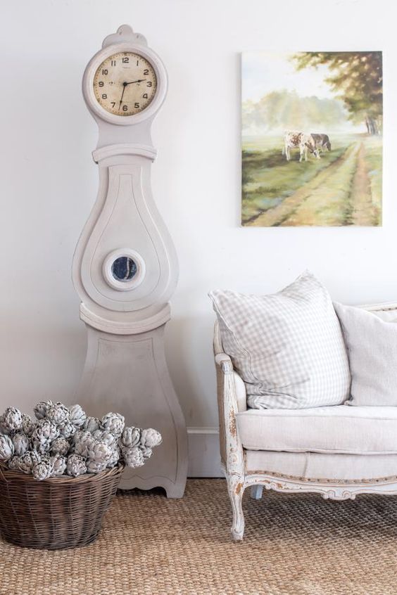 a lovely farmhouse space with a jute rug, a whitewashed vintage sofa with neutral pillows, a whitewashed grandfather clock, a basket with whitewashed pinecones