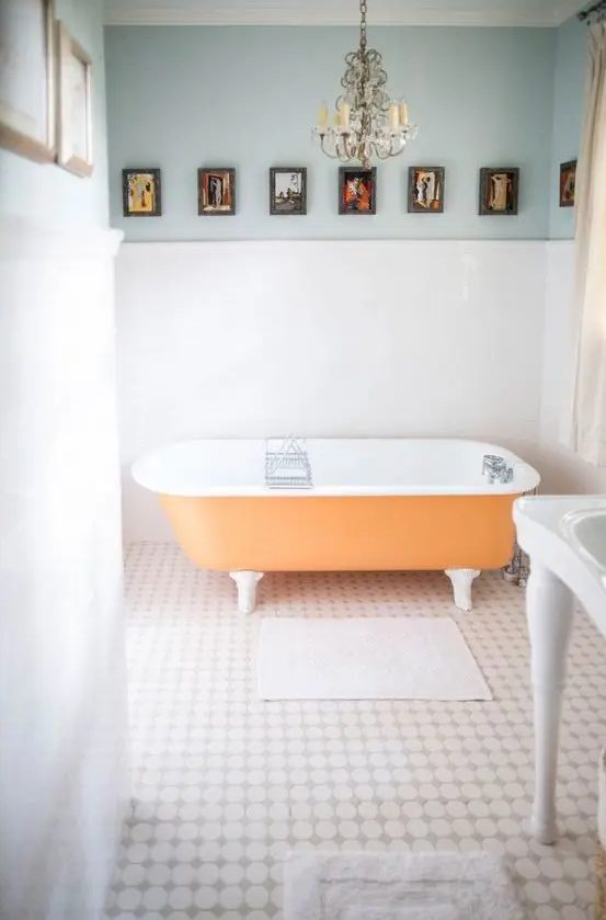 a light filled bathroom with light blue walls, an orange clawfoot bathtub, a crystal chandelier and a vintage gallery wall