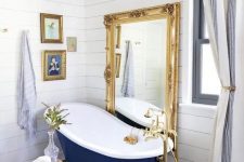 19 a refined bathroom with white shiplap, a navy clawfoot bathtub, a floor mirror in a gold frame, a crystal chandelier and artworks